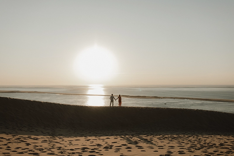 Un couple face à l'océan, se tient la main sur la dune du pilat au coucher du soleil.