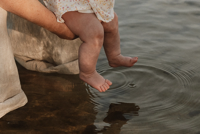 Une maman tient dans ses bras son bébé et se rapproche de l'eau pour qu'il la touche dans ses pieds.
