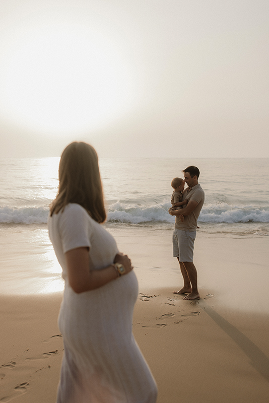 Une femme enceinte regarde son mari qui portent leur fils dans ses bras.